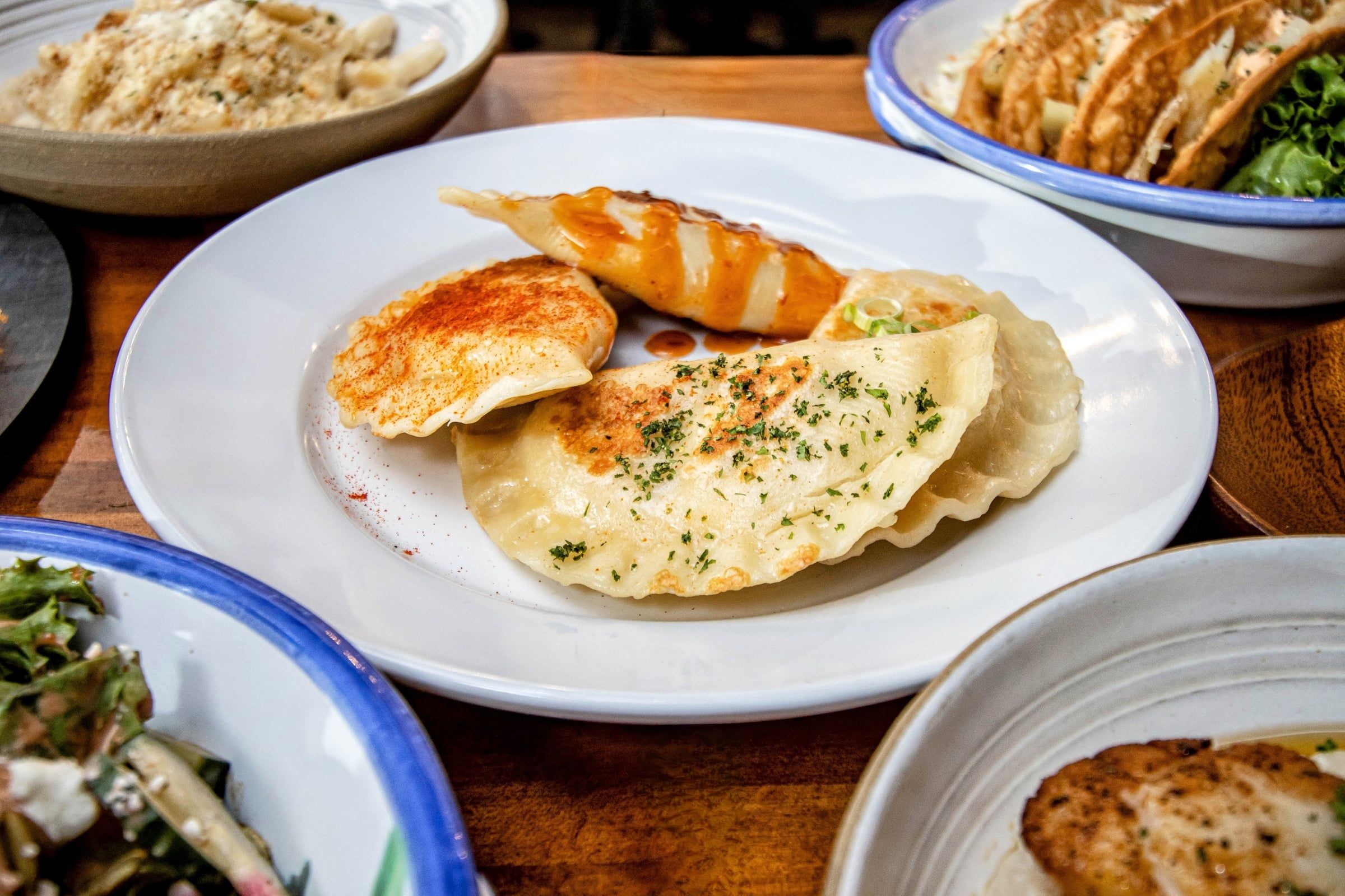 A plate of pan-fried pierogi with herbs and a drizzle of sauce, surrounded by other dishes