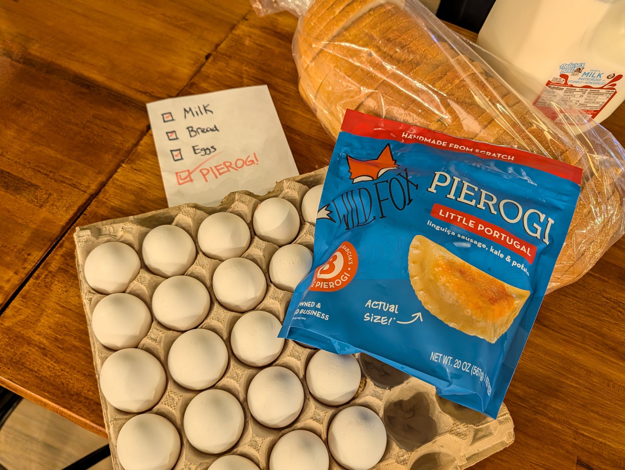 A bag of Little Portugal pierogi on a table with eggs, bread, and a shopping list: milk, bread, eggs, pierogi — with pierogi checked off